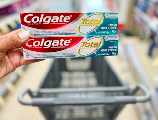 hand holding two boxes of Colgate Total toothpaste above shopping cart