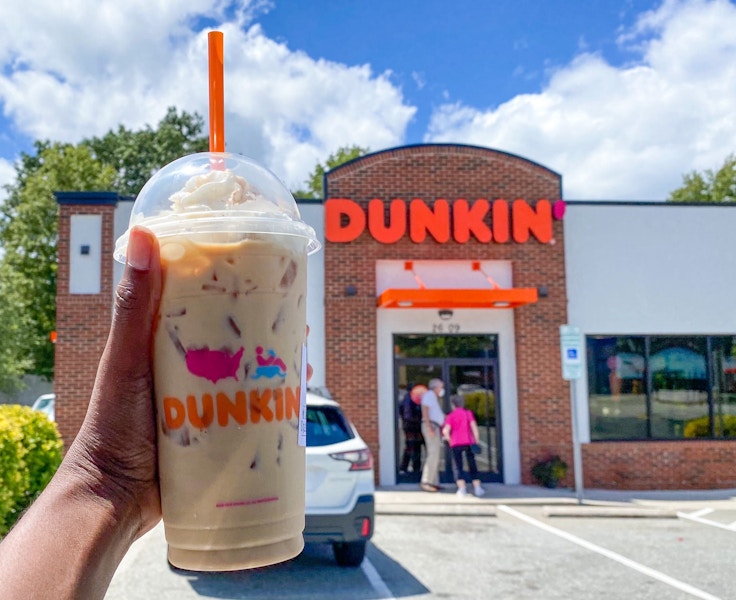 Someone holding the pumpkin spice latte in front of the dunkin' donuts store front.