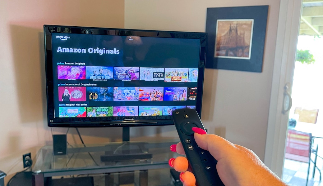 A person's hand holding an Amazon Fire remote, aiming it at a TV displaying Prime Video's Amazon Originals.