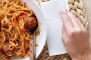 A plate of spaghetti and meatballs on a straw-woven placemat. A hand lays a Vanity Fair Extra Absorbent Napkin beside it.