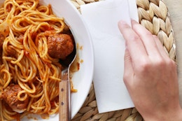 A plate of spaghetti and meatballs on a straw-woven placemat. A hand lays a Vanity Fair Extra Absorbent Napkin beside it.