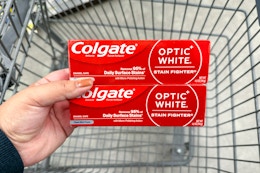 hand holding two tubes of colgate toothpaste over a shopping cart