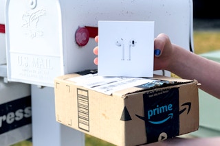 A person holding a box of Apple Airpods and an Amazon Prime delivery box near a mailbox.