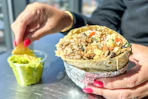 a person holding up a burrito and dipping a chip into guacamole 
