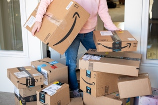 A woman picking up Amazon packages from a pile of Amazon boxes on a front doorstep.
