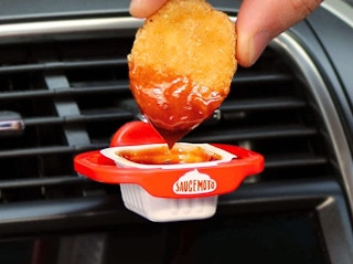 A person's hand dipping a chicken nugget into sauce being held by a Saucemoto dip clip attached to the car's A/C vent.