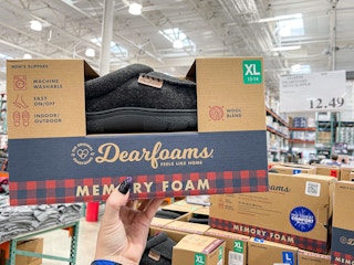 A person's hand holding up a box of Dearfoams memory foam slippers inside Costco.