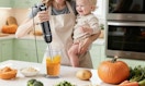 a mom holding a baby while using an immersion blender in a cup 