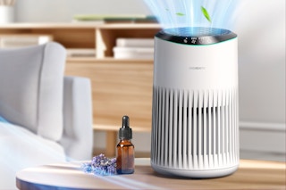 An air purifier sits on a table next to a sofa with a bottle of essential oil beside it.