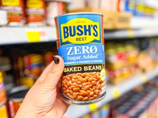 A hand holding a 27.5 ounce can of Bush's Zero Sugar Added baked beans in front of the bean aisle shelf at Walmart