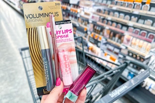 hand holding l'oreal mascara, maybelline lip makeup, and a lip liner in front of a walgreens cart in the makeup aisle