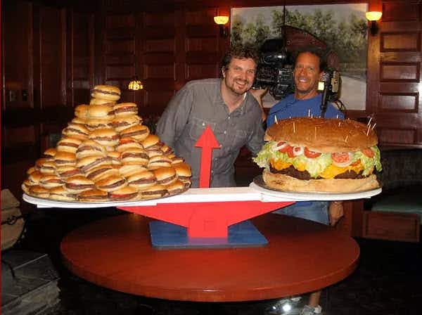 Two people standing behind a table with a large scale with an enormous burger on one side of the scale and a pile of smaller burgers on t...
