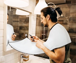 a man shaving over a beard bib over a bathroom sink