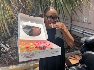 A woman eating a Mother's Day doughnut from Krispy Kreme, holding up a box of more doughnuts