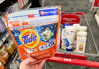 hand holding Tide pods in front of shopping cart with Head & Shoulder and Pantene hair care inside