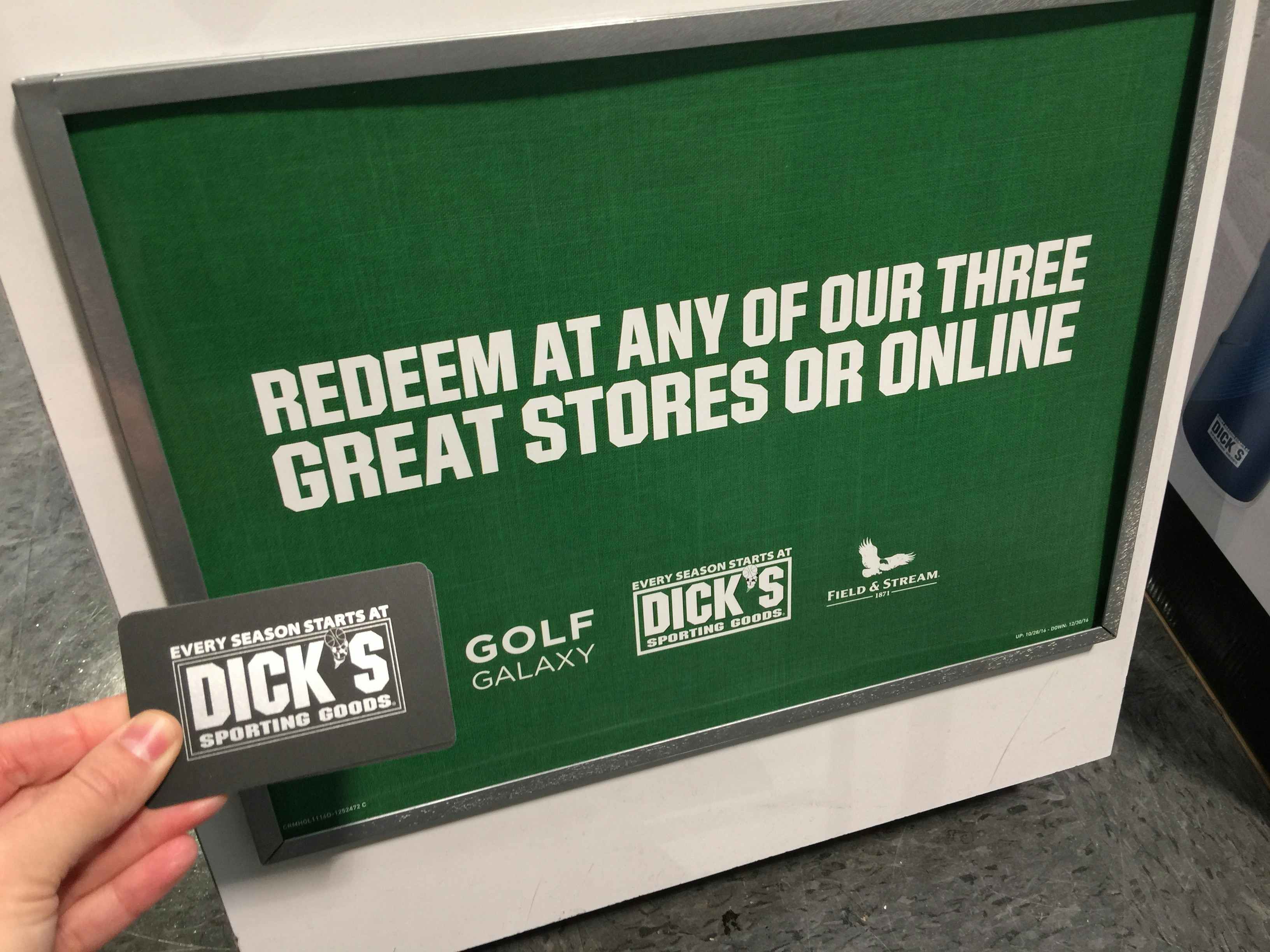 A person's hand holding a Dick's Sporting Goods gift card in front of a sign that reads "Redeem at any of our three great stores or onlin...
