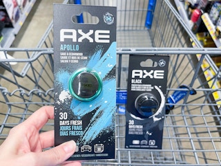 An Axe car freshener held out by hand in front of a cart with another axe car air freshener sitting in it.