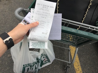 A person's hand holding a Dick's Sporting Goods bag and receipt, pushing a Dick's shopping cart.