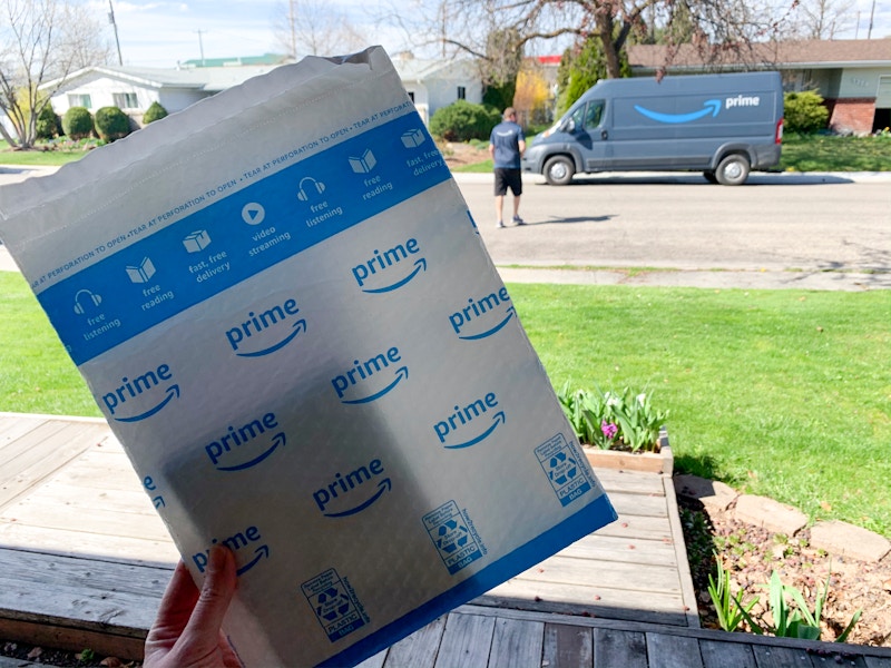 An amazon delivery package held up with a truck and delivery driver in the background.