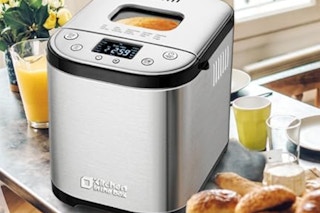 a bread maker machine on a counter with breakfast food and a glass orange juice