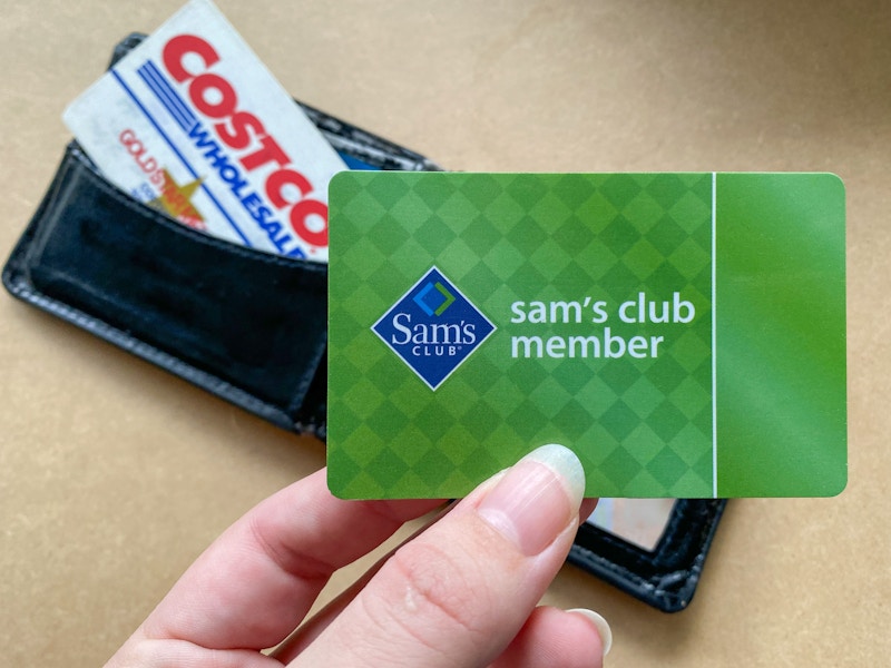 A person's hand holding a Sam's Club member card over a wallet with a Costco card in it.