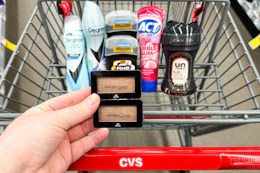person holding maybelline eyeshadow over a cart with degree deodorant, axe deodorant, act toothpaste, and downy unstoppables