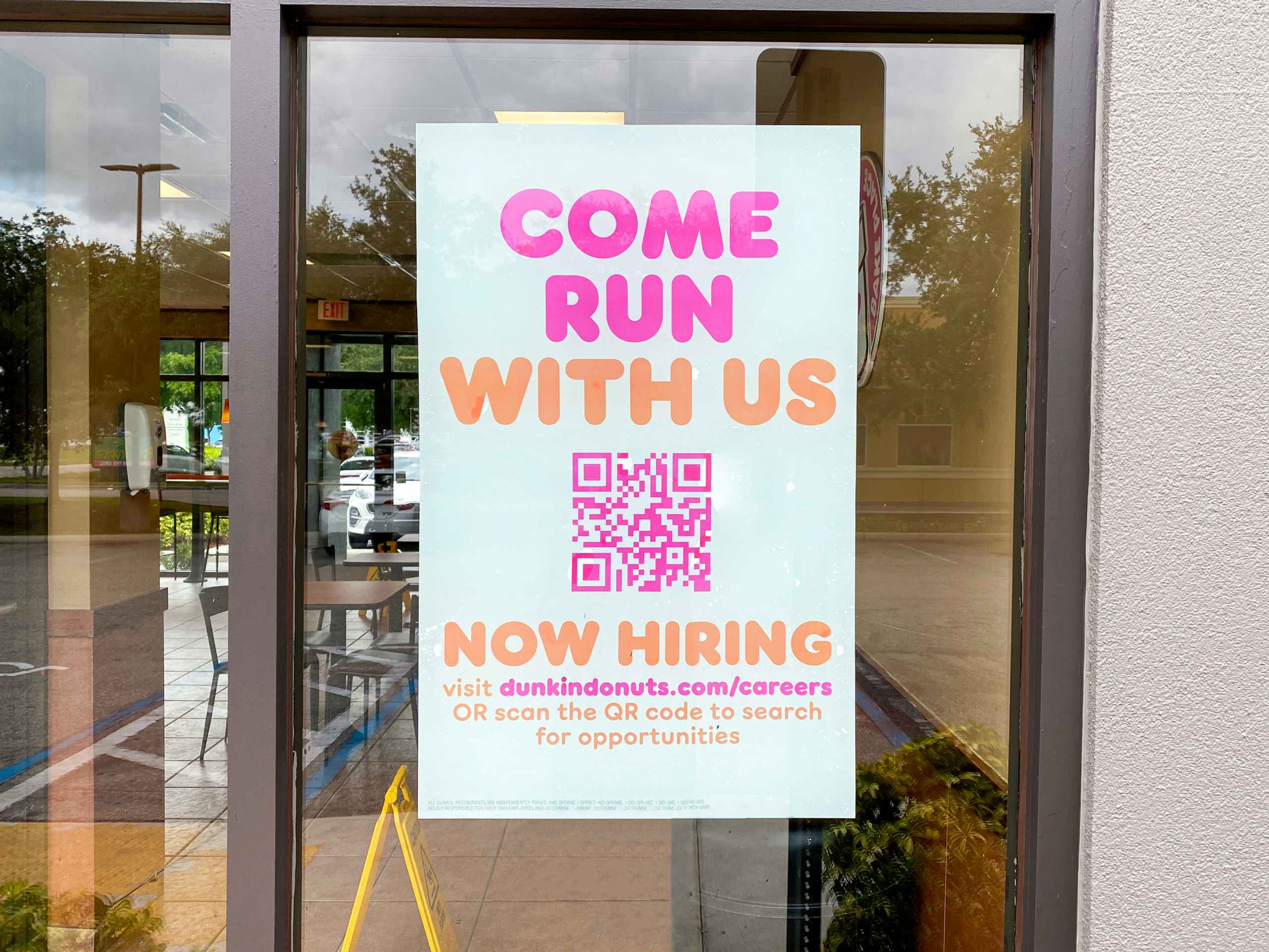 A sign in the window at Dunkin that says "Come run with us" with a QR code to apply for a position at Dunkin.
