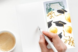 a person writing in a card with money that has grad caps and balloons on it