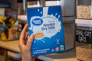 A person holding a box of Instant nonfat dry milk next to a shelf in a pantry