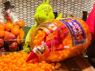 elf on the shelf doll inside a huge jar of cheese puffs with the puffs spilling out on counter and a grinch doll holding the container