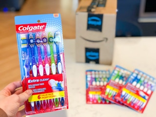 A hand holding a pack of Colgate toothbrushes in front of an Amazon delivery box.