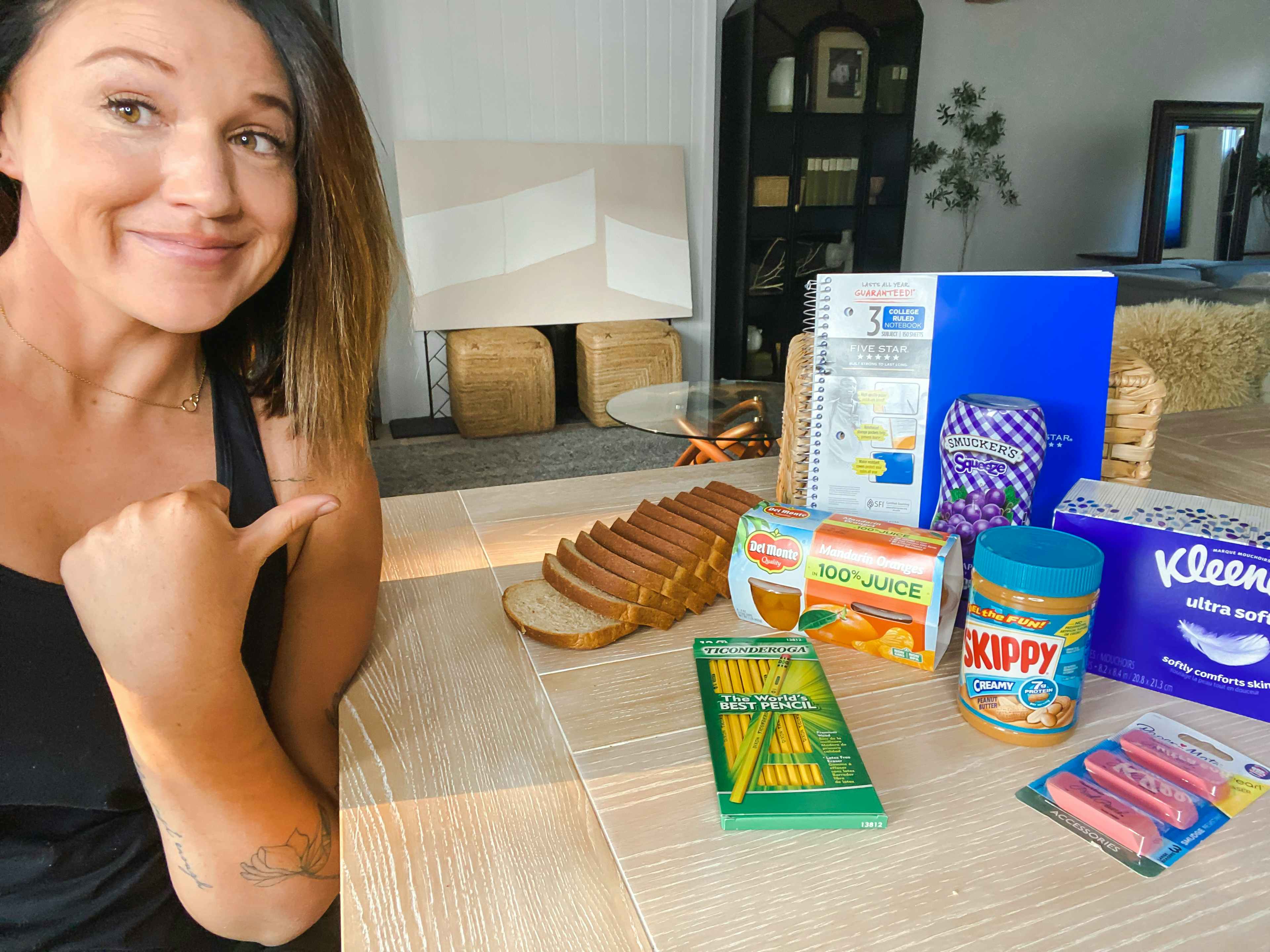 Woman sitting and pointing her thumb at some school supplies, bread, peanut butter, jelly, and a box of Kleenex on the table next to her.