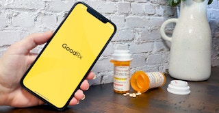 Someone holding a cell phone displaying the GoodRx app opening screen next to some prescription medications on a table