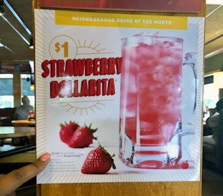 A sign for Applebee's Neighborhood Drink of the Month, advertising the $1 Strawberry Dollarita, with a person's hand pointing to the text...
