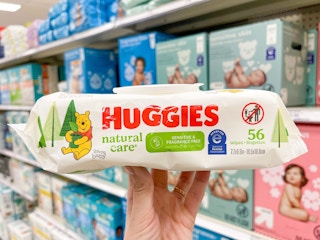 A package of Huggies wipes held out by hand in front of diapers sitting on a store shelf.