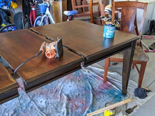 A dinning table with stain and a sander on top of it.