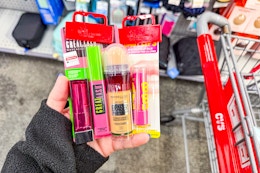 person holding maybelline lipstick, mascara, foundation, and baby lips by a cart