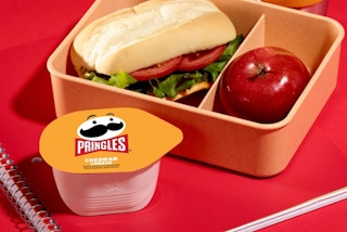 boxed lunch with sandwich, apple, and pringles stacks on a red notebook.