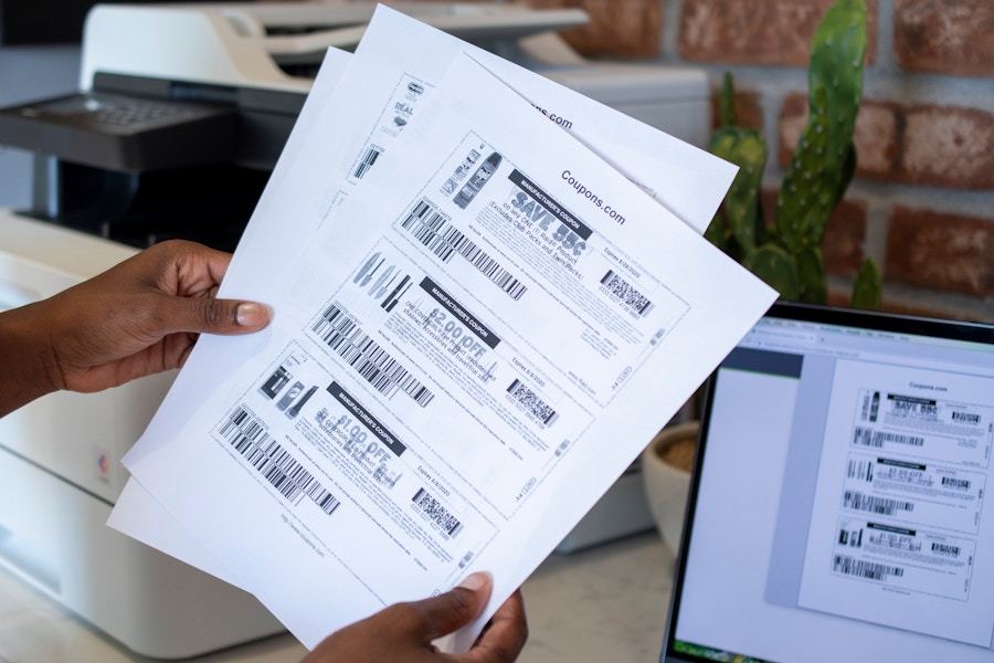 A person holding black and white printed coupons near a printer and computer.