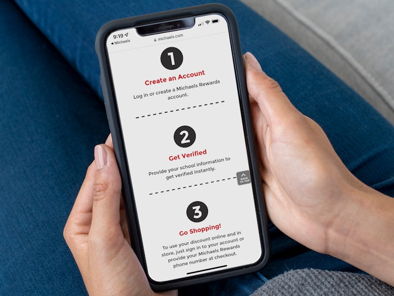 A person holding an iPhone displaying the instructions for Teacher Verification on the Michaels website.