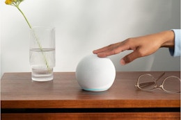 A hand taps a white Amazon Echo Dot on a bedside table