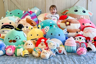 A child sitting on a bed covered in a huge pile of different sized Squishmallows.