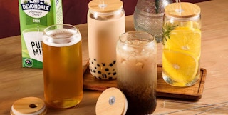 a set of 4 glass beverage cups with bamboo lids and glass straws filled with drinks on a wooden table