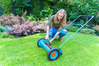 A teenage girl adjusting the settings on a manual push lawn mower on someone's lawn.