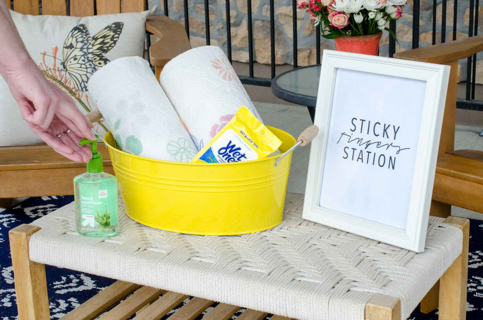 Person reaching for hand sanitizer next to a bucket with rolls of paper towels and Wet Ones on a table with a sign that reads, "Sticky fi...