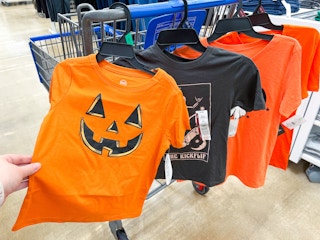 hand holding a halloween tee with others hanging on walmart cart
