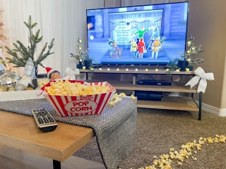 elf on the shelf doll laying inside a tray of popcorn in front of a tv with elf movie on screen