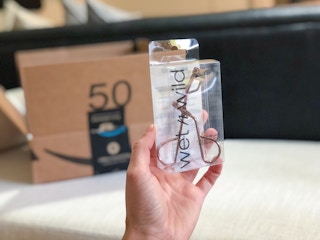A hand holding a Wet n Wild eyelash curler in front of a delivery box.