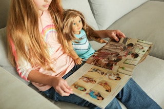 A girl, sitting on a couch with her American girl doll, reading the American Girl catalogue.