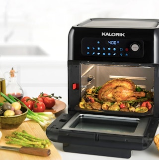 An air fryer with a roasted chicken and vegetables inside on a counter surrounded by plates of food.